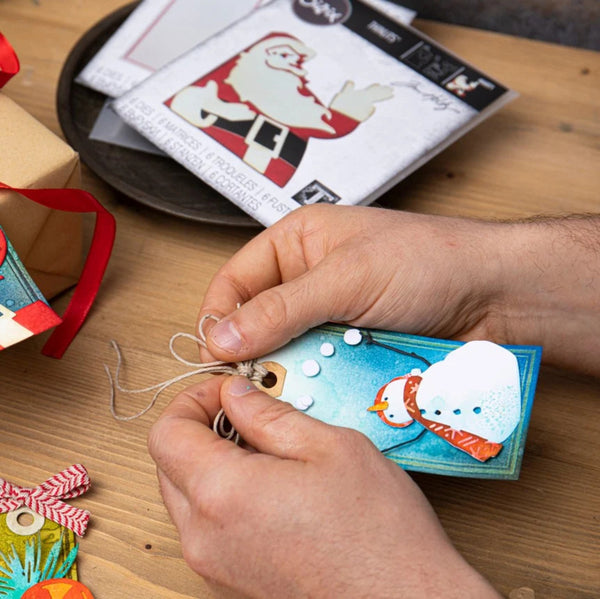 A close-up of hands crafting a Christmas ornament tag with a snowman design, working on a wooden table with a package of Santa-themed embellishments in the background.