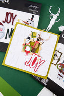 A handmade Christmas greeting card with layered white and olive green borders, featuring a snowman, reindeer, wreath, and red 'JOY' lettering, styled on a green background with Thintlits dies packaging.