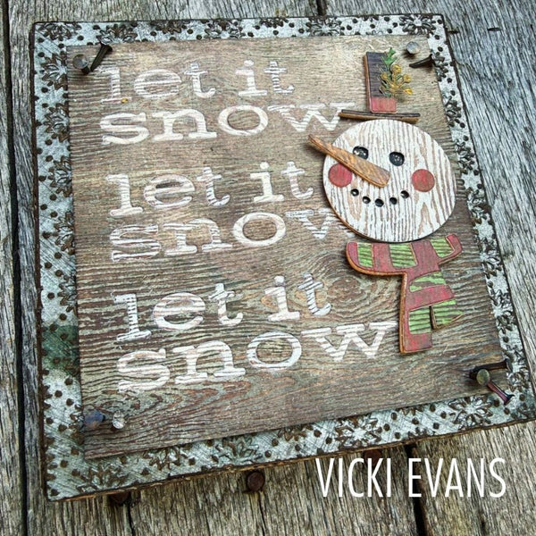 A rustic decorative wooden sign with the phrase "let it snow let it snow let it snow" in distressed white lettering, featuring a raised snowman face and scarf on the right. The sign has a distressed metal border and is photographed on weathered wood planks.
