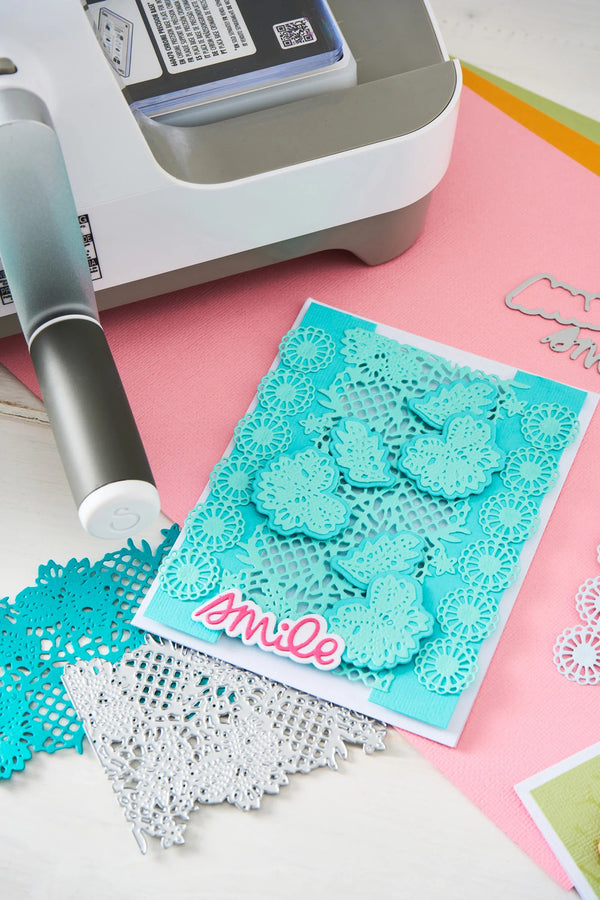 Card with decorative cutouts and 'smile' text on a pink surface with crafting tools.