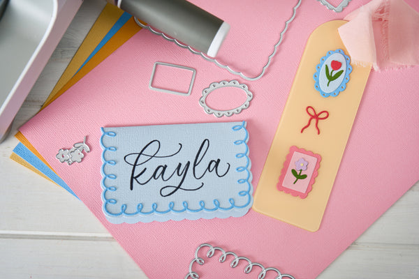 A flat-lay of crafting supplies on a pink surface, featuring a central handmade card with the name 'Kayla' in black script inside a light blue scalloped border, surrounded by metal embossing dies, colored papers, and decorative tags.
