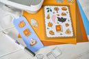 A flat-lay of a crafting kit featuring a blue die-cut tag with pumpkin and autumn-themed stickers, a sheet of layered autumn stickers on brown cardstock, orange and blue cardstock sheets, a white embosser tool, and metal cutting dies on a white wood background.