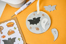 A Halloween-themed metal ornament set displayed on a textured orange background, featuring a round silver ornament with black and white bat and ghost cutouts, along with individual bat and ghost cutouts and a sticker sheet.