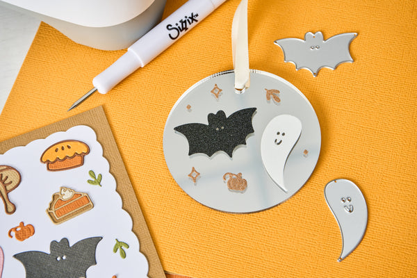 A Halloween-themed metal ornament set displayed on a textured orange background, featuring a round silver ornament with black and white bat and ghost cutouts, along with individual bat and ghost cutouts and a sticker sheet.