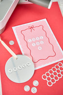 A pink and white card with the words 'LOVE YOU' made from small white letter charms, displayed on a red background with crafting tools and metal rings around it.