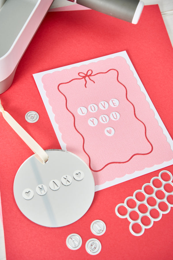 A pink and white card with the words 'LOVE YOU' made from small white letter charms, displayed on a red background with crafting tools and metal rings around it.