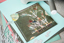 A handmade greeting card featuring a tree with birds and the text 'TREE AS A BIRD' on a dark olive background, styled on a light blue cardstock with crafting tools and paper around it.