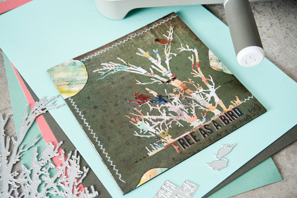 A handmade greeting card featuring a tree with birds and the text 'TREE AS A BIRD' on a dark olive background, styled on a light blue cardstock with crafting tools and paper around it.
