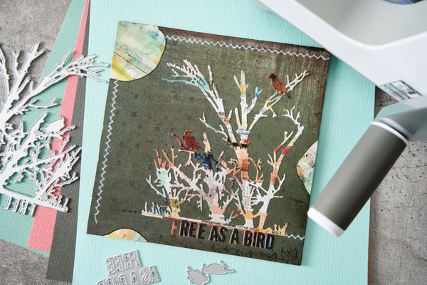 A handmade bird-themed scrapbook card featuring distressed brown wood, white-painted tree branches, small birds, and the text 'FREE AS A BIRD', styled on a light blue mat with crafting tools and dies around it.