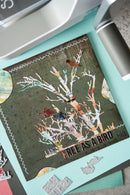 A close-up of a handmade scrapbook card featuring a tree with birds perched on its branches, layered over a textured dark green background. The card is displayed on a light teal crafting mat with die-cut paper elements and a metallic die visible in the foreground.