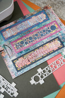 A close-up of a crafting layout showing three colorful rectangular patterned strips arranged on a dark gray mat, with white metal die-cutting templates placed around them on pink, teal, and orange paper sheets.