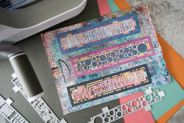 A handmade mixed-media art card featuring three horizontal bands of colorful geometric patterns, displayed on a dark gray surface alongside crafting tools and colorful paper sheets.