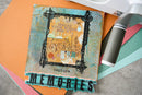 A square mixed-media art card or scrapbook piece featuring a distressed turquoise and orange design with the words 'SPREAD YOUR WINGS AND FLY' and 'capture MEMORIES' at the bottom, styled on top of layered colored paper sheets with a white tool partially visible.