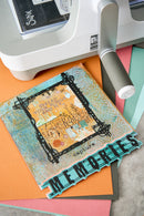 A close-up of a handmade scrapbook card featuring a vintage-style frame design with the words 'Capture Memories' and a turquoise 'Memories' banner, styled on top of orange and pink cardstock with a white crafting machine partially visible in the background.