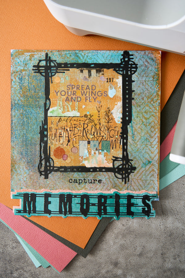 A square mixed-media art card or scrapbook paper featuring a distressed teal and gold design with the words 'SPREAD YOUR WINGS AND FLY' and 'capture MEMORIES' in layered black and teal text, styled on top of other colorful paper sheets.