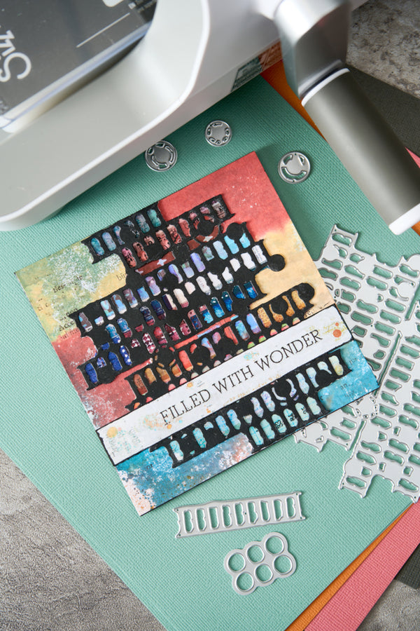 A close-up of a handmade layered card featuring a colorful abstract cityscape design with the text 'FILLED WITH WONDER'. The card is displayed on a mint green cardstock mat alongside metal die-cutting and embossing tools, including a white die and a silver embosser, on a gray textured surface.