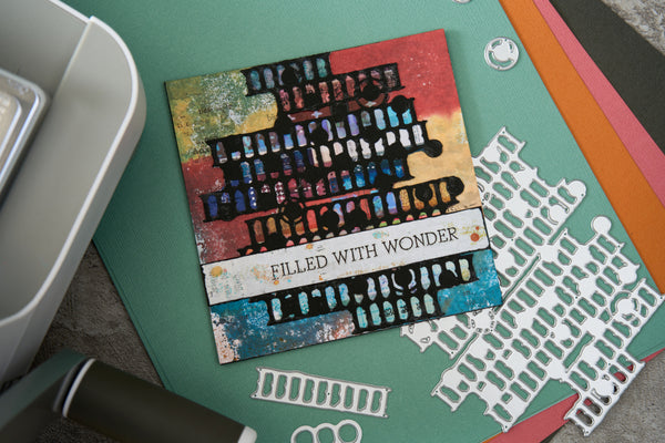 A square handmade card with a colorful abstract design and the text 'FILLED WITH WONDER' lies on a green crafting mat, surrounded by die-cut paper pieces, a white tray, and other craft supplies.