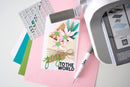 A close-up flat lay of a handmade greeting card featuring pink flowers, green leaves, and the text 'J'YOU TO THE WORLD' in green and black. Surrounding the card are crafting supplies including a white die-cut pen with a Skripa logo, a white cutting machine, colorful cardstock sheets, and a sticker sheet on a light gray surface.