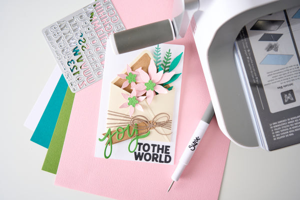 A close-up flat lay of a handmade greeting card featuring pink flowers, green leaves, and the text 'J'YOU TO THE WORLD' in green and black. Surrounding the card are crafting supplies including a white die-cut pen with a Skripa logo, a white cutting machine, colorful cardstock sheets, and a sticker sheet on a light gray surface.