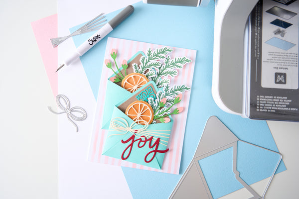 A handmade layered card with teal envelope details, green foliage, orange slices, and the word 'joy' in red script, styled on a light blue and white surface with crafting tools nearby.