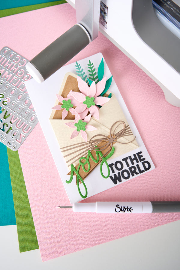 A handmade greeting card with layered paper flowers, green leaves, and the text 'you TO THE WORLD' in green and black, styled on a pink surface with crafting tools nearby.