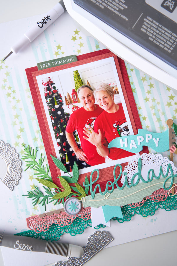 A close-up flat lay of a scrapbook or photo layout featuring a printed photo of two people in red Christmas shirts, surrounded by layered paper elements including a teal 'happY holiday' banner, green paper foliage, lace borders, and crafting tools with Sizzl and Silhouette branding.