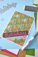 A handmade holiday greeting card with green script lettering, blue snowflakes, and a pink 'holiday' banner, styled on a light blue background with craft tools and paper.