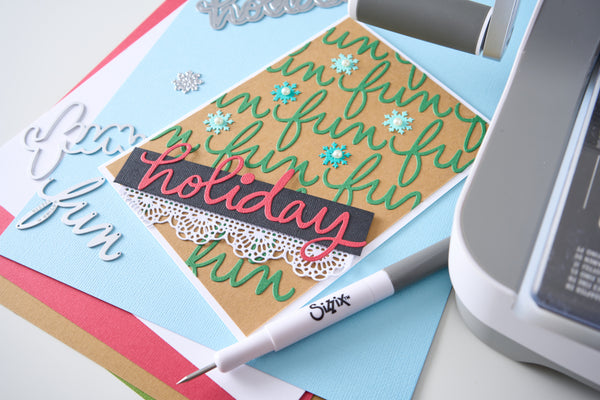 A handmade holiday greeting card with 'fun holiday fun' in green and pink script, decorated with blue snowflakes and a white lace border, shown alongside crafting tools and papers.