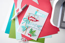 A handmade Christmas greeting card with layered paper cutouts and the text 'HAVE A MERRY LITTLE CHRISTMAS' in light blue, red, white, and green, styled on top of colored cardstock sheets with a Dymo pen and a die-cutting machine visible around it.
