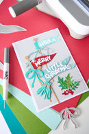 A handmade layered Christmas greeting card with 'HAVE A MERRY LITTLE CHRISTMAS' in decorative paper cutouts, styled on a flat-lay craft setup with colored cardstock and crafting tools.