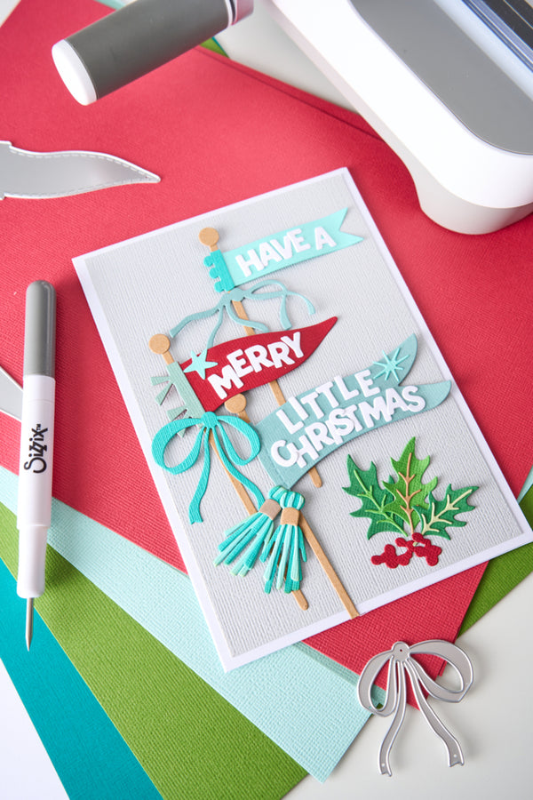 A handmade layered Christmas greeting card with 'HAVE A MERRY LITTLE CHRISTMAS' in decorative paper cutouts, styled on a flat-lay craft setup with colored cardstock and crafting tools.