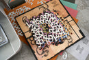 A square craft card or scrapbook base with a distressed tan background, black border, and black decorative flourishes, overlaid with a dense layer of white, black, orange, purple, and blue numerals reading 1 through 9. The card is photographed on top of other colorful paper sheets and craft materials on a light gray surface.