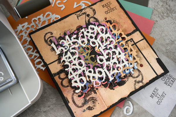 A square craft card or scrapbook base with a distressed tan background, black border, and black decorative flourishes, overlaid with a dense layer of white, black, orange, purple, and blue numerals reading 1 through 9. The card is photographed on top of other colorful paper sheets and craft materials on a light gray surface.