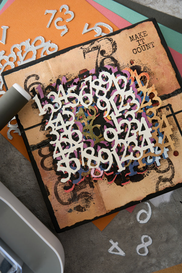 A square craft die-cut paper sheet featuring a distressed tan background with black ornamental swirls, scattered white numbers 1 through 9, and the words 'MAKE IT COUNT' in the top right. The sheet is photographed on top of colorful paper stacks and other craft dies, with a die-cutting machine partially visible at the edges.