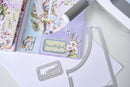 A close-up flat lay of a crafting kit showing a lavender card with floral illustrations and the word 'beautiful,' alongside metal die-cutting or embossing tools and patterned paper sheets on a white surface.