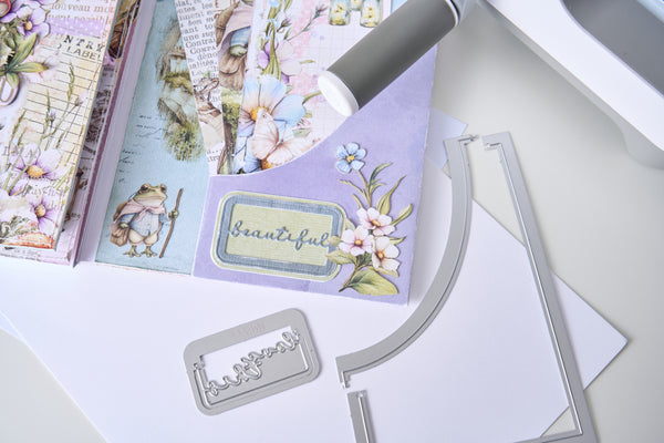 A close-up flat lay of a crafting kit showing a lavender card with floral illustrations and the word 'beautiful,' alongside metal die-cutting or embossing tools and patterned paper sheets on a white surface.