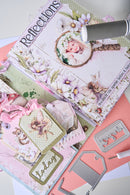 A styled flat-lay of scrapbooking and crafting supplies, featuring a decorated journal with a fairy photo, pink ribbons, paper tags reading 'today' and 'Reflections', metal die-cut stamps, and a silver heat gun on a pastel background.