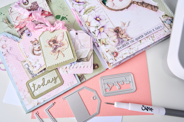 A styled flat-lay of scrapbooking and crafting supplies, including layered paper albums with fairy and floral designs, metal die-cutting templates, a pink pen, and a white cutting machine on a light gray surface.