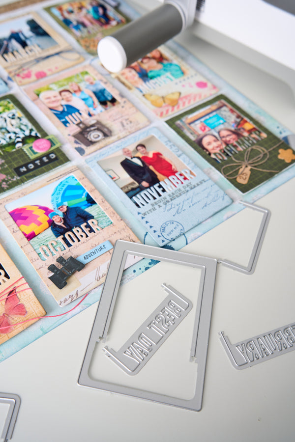 A close-up of a crafting workspace showing a metal die-cut frame with the word 'MERRY' and other silver metal frame dies scattered on a white surface, alongside sample photo cards and a white rotary cutter.