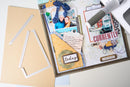 A styled flat-lay of scrapbook crafting supplies on a white surface, including a large beige cardstock sheet, white frame cutouts, a decorated layered paper page with photos and text, a gray and white pen, and a small blue ribbon.