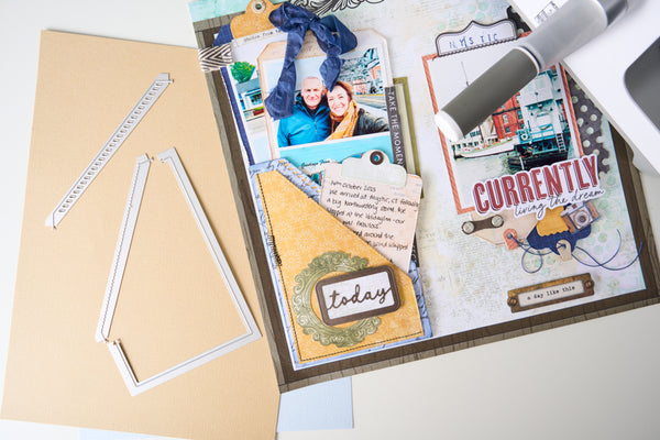A styled flat-lay of scrapbook crafting supplies on a white surface, including a large beige cardstock sheet, white frame cutouts, a decorated layered paper page with photos and text, a gray and white pen, and a small blue ribbon.