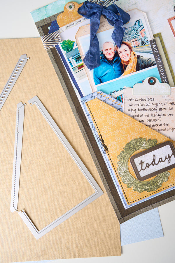 A close-up of a scrapbook or card-making craft kit showing layered paper elements, a photo of two people, decorative tags, a blue ribbon, and metal die-cut frames on a beige background.