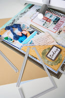 A close-up of a scrapbook or craft layout featuring layered paper elements, a small photo of two people, decorative text labels, and metal frame cutters on a light surface.