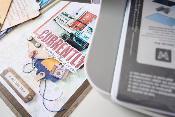 A close-up of a scrapbook or craft paper layout featuring layered paper elements, a camera illustration, blue ribbon, and the words 'Currently living your dream.' A printer or scanner is partially visible on the right side of the frame.