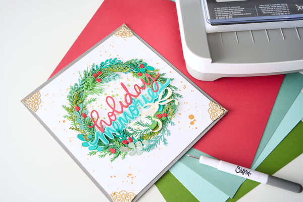 A square handmade greeting card with a festive wreath design and the words 'holiday memories' in layered script, placed on top of red and green cardstock with a white die-cutting or stamping machine partially visible in the upper right.