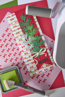 A festive handmade Christmas card featuring green pine branches, a red background, a white and green striped bow, and the word 'NOEL' in black. The card is styled on a crafting surface with other paper tools and ink pads.