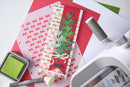 A handmade Christmas greeting card with green pine branches, a red background, and a cream bow, displayed on a crafting mat with other craft tools including an ink pad and a die-cutting machine.