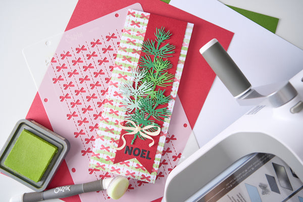 A handmade Christmas greeting card with green pine branches, a red background, and a cream bow, displayed on a crafting mat with other craft tools including an ink pad and a die-cutting machine.
