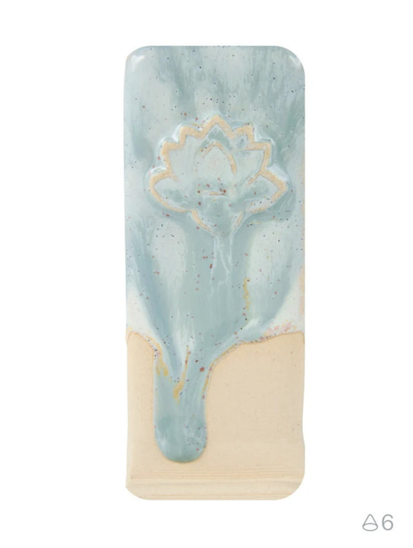 A rectangular ceramic tile or plaque with a glossy light blue and cream drip design, featuring a raised gold lotus flower motif near the top, shown on a plain white background.
