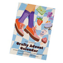 Spellbinders Crafty Advent Calendar book cover shown at an angle on a plain white background. The cover features a whimsical illustration of a person's legs in purple socks and pink shoes, alongside pastel drawings of an envelope, strawberry, cake, flower, and heart on a blue-and-white checkered pattern.